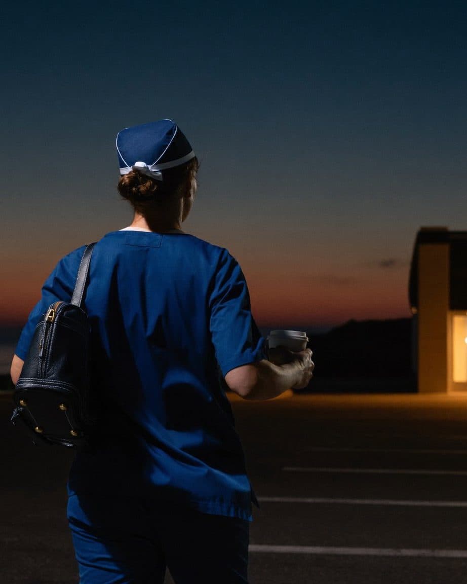 Pre-dawn at a hospital entrance: a nurse in navy scrubs with a shoulder bag and travel mug walks toward the warm light of the doors to start an early shift.