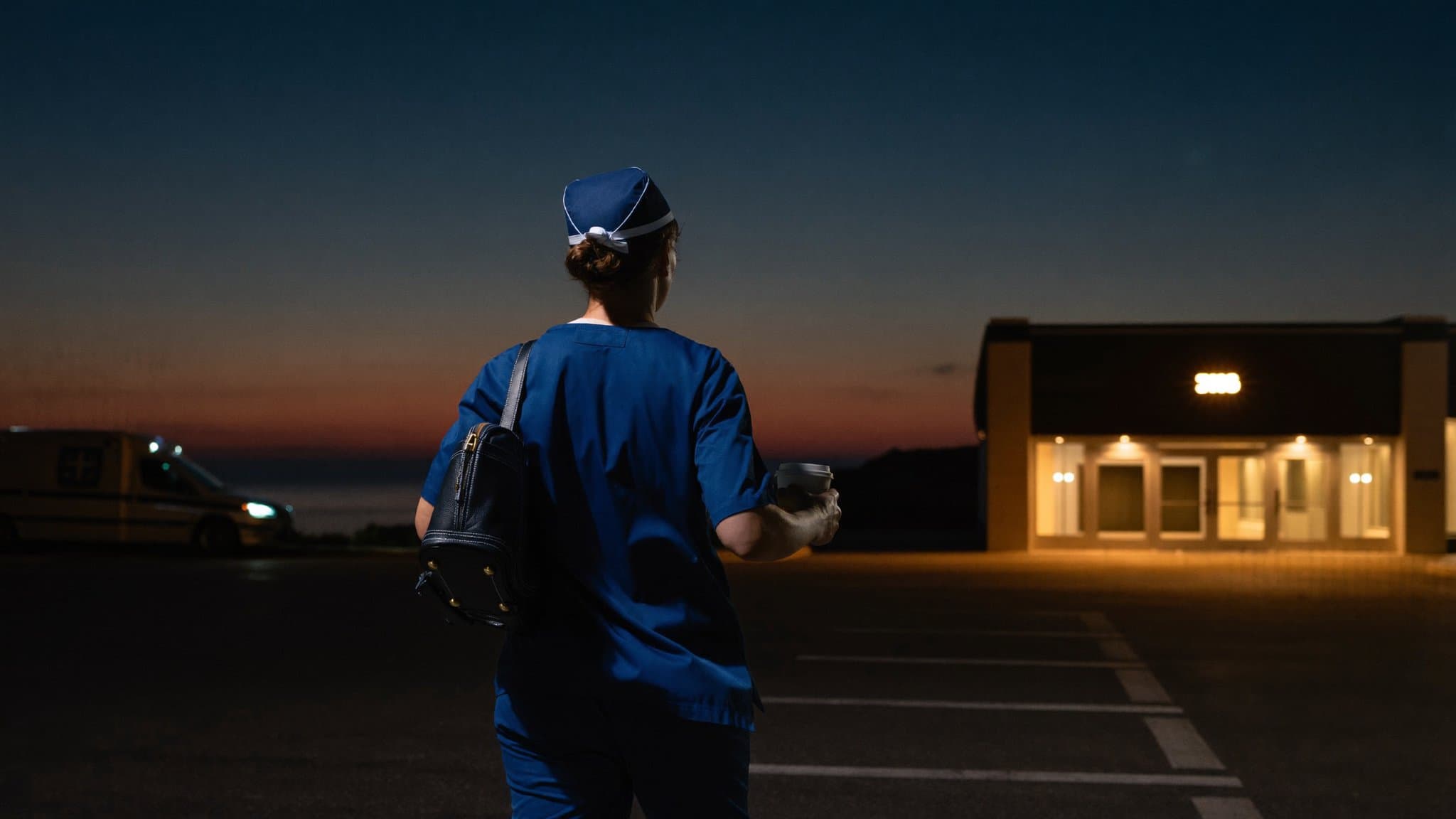 Pre-dawn at a hospital entrance: a nurse in navy scrubs with a shoulder bag and travel mug walks toward the warm light of the doors to start an early shift.